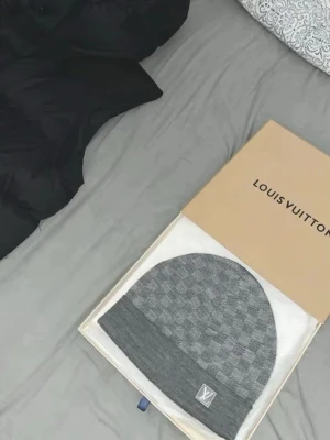 Grå Louis Vuitton mössa - Säljer en snygg grå mössa från Louis Vuitton med diskret rutmönster och LV-logga framtill. Mössan är stickad och har en bredare kant nedtill för extra stil och komfort. Perfekt accessoar för dig som gillar exklusiva märken och klassisk design.