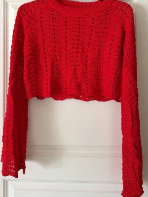 Röd virkad cropped tröja - Trendig röd stickad cropped tröja med hålmönster och långa ärmar. Tröjan har en rund halsringning och vågig kant nertill. Perfekt för att styla med höga jeans eller kjol. Materialet känns lätt och luftigt. Märke LXA. Helt oanvänd