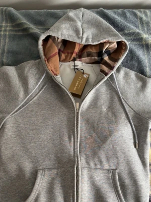 Grå zip hoodie från Burberry - Snygg grå zip hoodie från Burberry med klassiskt rutigt foder i huvan och dragsko. Hoodien har fickor framtill och en diskret broderad logga på bröstet. Perfekt för dig som gillar stilrena och exklusiva plagg. Materialet känns mjukt och bekvämt.