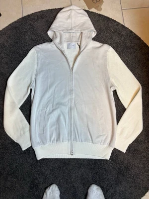 Gran Sasso cardigan - Säljer en stilren vit cardigan från Gran Sasso med dragkedja framtill och två fickor. Hoodien har ribbade muddar och ärmar i stickad struktur för extra detalj. Perfekt för en clean och avslappnad look.