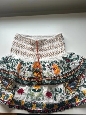 Vit broderad volangkjol med snörning - Supersöt vit kjol med broderade blommor i grönt, gult och rött. Kjolen har volanger, färgglad snörning i midjan och elastisk smock upptill. Perfekt för dig som gillar boho-stil och detaljerade mönster.