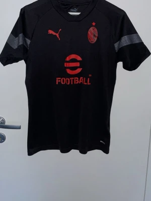 Svart AC Milan matchtröja Puma - Snygg svart AC Milan matchtröja från Puma med röda detaljer och klubbmärke på bröstet. Tröjan har korta ärmar med gråa ränder och är tillverkad i ett lätt, andningsbart material som passar perfekt till träning eller match.