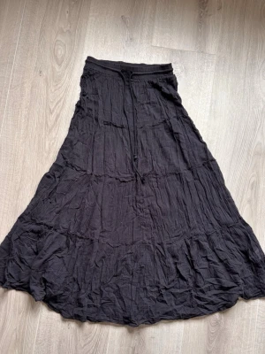 Svart långkjol med snörning - Svart långkjol i luftig bomull med resår och snörning i midjan. Kjolen har en avslappnad passform och flera volangliknande sömmar som ger ett bohemiskt intryck. Perfekt för dig som gillar en enkel men snygg stil.