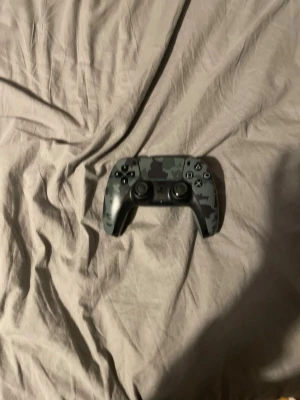 Sony DualSense camo handkontroll PS5 - Snygg DualSense handkontroll till PS5 med grått och svart camomönster på framsidan och helgrå baksida. Ergonomisk design med klassiska PlayStation-knappar och två analoga spakar. Perfekt för gaming och passar alla som vill ha en unik look på sin kontroll. PRISET KAN DISKUTERAS.