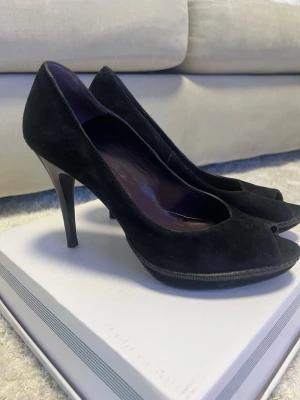 Svarta peep toe pumps från Novita - Snygga svarta pumps från Novita i mocka med peep toe och hög klack. Insidan är fodrad i mörklila skinn och sulan har en diskret platå framtill. Perfekta för dig som vill ha en klassisk och elegant look.