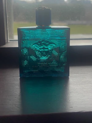 Versace Eros Eau de Toilette - Versace Eros Eau de Toilette i en snygg turkos glasflaska med Medusa-huvud i relief och guldfärgade detaljer. Flaskan har en fyrkantig form och känns riktigt lyxig och modern. Perfekt för dig som gillar exklusiva dofter och vill ha en parfym som sticker ut i badrumshyllan.