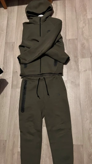 Olivgrön Nike tracksuit med huva - Säljer en olivgrön hoodie från Nike med hel dragkedja framtill och två fickor med svarta dragkedjor. Hoodien har en klassisk huva och diskret Nike-logga på bröstet. Materialet är mjukt och stretchigt, perfekt för en avslappnad streetwear-look.