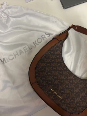 Brun axelremsväska från Michael Kors - Snygg handväska från Michael Kors i brunt konstläder med klassiskt MK-mönster över hela väskan. Väskan har guldfärgade detaljer, dragkedja upptill och en bred axelrem. Lite skavanker på insidan, se sista bild