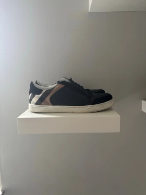Burberry svart/vit låg sneaker - Säljer ett par snygga Burberry sneakers i svart med klassiskt rutmönster i beige, vitt och svart på sidan. Skorna har vita sulor, svarta skosnören och detaljer i mocka och läder. Perfekta för dig som vill ha en stilren och trendig look.