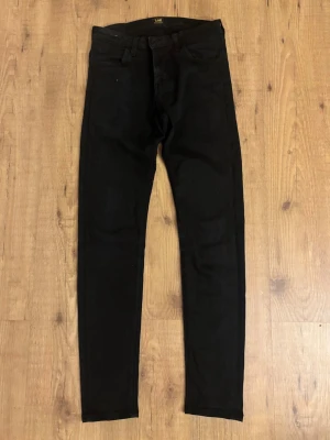 Svarta skinny jeans Malone från Lee - Säljer ett par svarta skinny jeans Malone från Lee med klassisk femficksmodell och hög midja. Jeansen har smal passform och är tillverkade i ett mjukt denimtyg som sitter snyggt. Perfekta till en avslappnad streetwear-look.