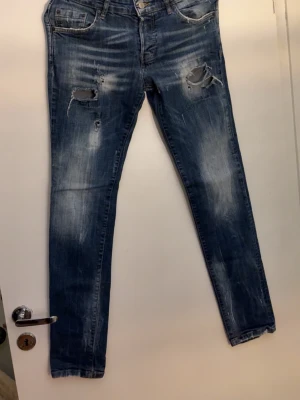 Blå slitna skinny jeans Dsquared - Säljer ett par blå skinny jeans med slitningar och hål på låren och knäna. Jeansen har en tvättad look med ljusare partier och klassisk femficksdesign. Perfekta för en avslappnad och trendig stil.