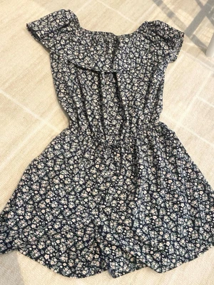 Blommig kortklänning från Name It - Söt klänning från Name It med mörkblå botten och små vita och orange blommor. Klänningen har volangdetalj vid axlarna och resår i midjan för en bekväm passform. Perfekt för varma dagar och enkel att styla. !Kjolen är inte helt lös utan har en liten delning för benen!