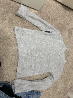 Ljusgrå stickad tröja - Mysig ljusgrå stickad tröja med rund hals och långa ärmar. Tröjan har en enkel, rak passform och ribbstickade muddar vid ärmslut och nederkant. Perfekt för lager på lager under kyliga dagar. Storlek S 