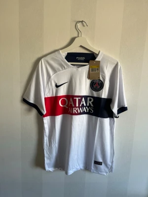 PSG Mbappé matchtröja vit Nike - Snygg vit PSG matchtröja från Nike med Mbappé och nummer 7 på ryggen. Tröjan har röda och marinblå detaljer, Qatar Airways sponsortryck och PSG-logga på bröstet. Tillverkad i lätt och ventilerande material, perfekt för fotboll eller träning.