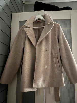 Beige kappa från RYVLS med bälte - Stilren beige kappa från RYVLS med dubbelknäppning och bred krage. Kappan har raka ärmar, två sidofickor och ett matchande bälte i midjan. Tillverkad i ett mjukt, ull-liknande material som ger en klassisk look.