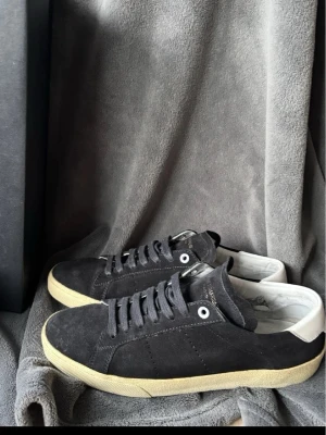 Svarta Saint Laurent sneakers i mocka - Säljer ett par svarta sneakers från Saint Laurent i mjuk mocka med vita detaljer och beige sula. Klassisk låg modell med snörning och diskret logga på insidan. Perfekta för dig som gillar stilrena och exklusiva sneakers.