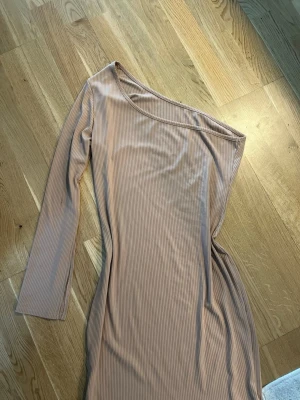 Beige oneshoulder ribbad klänning - Trendig beige oneshoulder klänning med lång ärm och ribbad struktur. Klänningen har en kroppsnära passform och är perfekt för dig som vill sticka ut med en stilren look. Materialet känns mjukt och följsamt mot kroppen.
