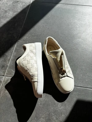 Beige sneakers från Michael Kors - Snygga beige sneakers från Michael Kors med guldfärgade dragkedjor och MK-logga på sidan. Skorna köptes på NK i Göteborg inför nyår 2025 då de användes och har ej används sedan dess! Nypris 1500kr! Buda gärna 