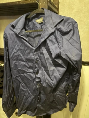 Marinblå skjorta med knappar - Klassisk marinblå skjorta från Dressmann med lång ärm och knappar framtill. Skjortan har en bred krage och är tillverkad i ett mjukt bomullsmaterial. Perfekt för dig som gillar stilrena och enkla plagg.