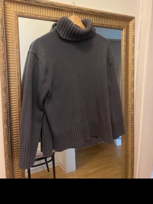 Mörkgrå polotröja i stickat - Mysig mörkgrå stickad polotröja med bred ribbstickad kant nedtill och vid ärmsluten. Tröjan har en hög, tjock polokrage och en avslappnad passform. Perfekt för kalla dagar och enkel att matcha med olika outfits.