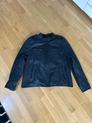 Svart skinnjacka från Levi's - Snygg svart skinnjacka från Levi's med fyra dragkedjeförsedda fickor och ståkrage med knapp. Insidan har rutigt foder i grått och rött. Perfekt för dig som vill ha en klassisk och stilren look.