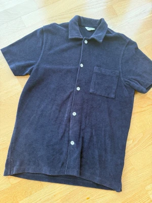 Mörkblå frottéskjorta med kort ärm från East West - Mjuk och avslappnad mörkblå skjorta i frotté med korta ärmar, klassisk krage och vita knappar framtill. Bröstficka på vänster sida ger en chill vibe. Perfekt för sommarens alla dagar.