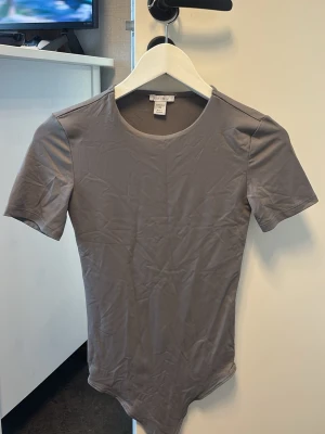 Grå kortärmad body från Cubus - En stilren grå body från New Yorker med korta ärmar och rund halsringning. Tillverkad i ett mjukt och stretchigt material som sitter skönt mot kroppen. Perfekt att styla med jeans eller kjol för en enkel och clean look. Aldrig använt behöver bara strykas 