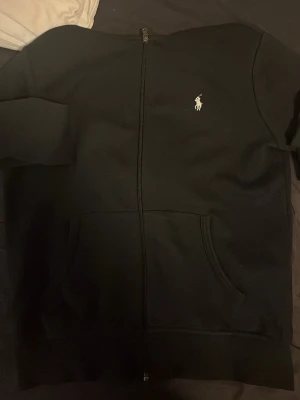 Svart hoodie från Polo Ralph Lauren - Svart hoodie med dragkedja från Polo Ralph Lauren. Klassisk design med vit broderad logga på bröstet, känguruficka framtill och huva med snörning. Perfekt för en chill och stilren look. Materialet känns mjukt och bekvämt.