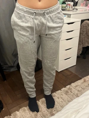 Grå mjukisbyxor från Nike - Säljer ett par ljusgrå mjukisbyxor från Nike i storlek XS. Byxorna har dragsko i midjan, muddar vid bensluten och är tillverkade i mjukt, bekvämt material. Perfekta för chill dagar eller träning. Klassisk Nike-logga på etiketten.