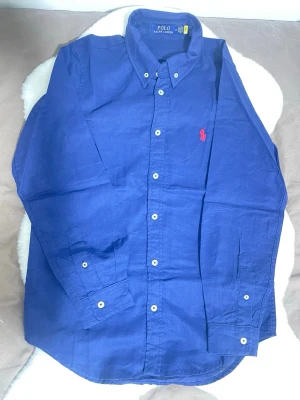 Blå linneskjorta från Polo Ralph Lauren - Säljer en klassisk marinblå skjorta från Polo Ralph Lauren med button-down krage och blå broderad logga på bröstet. Skjortan har långa ärmar och knäppning framtill. Den är helt ny och i bästa skick! Perfekt för sommaren☀️  Skick[10/10] storlek[M] pris[549]  Hör av dig vid intresse och mer info 🥂