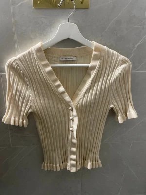 Ribbad beige kofta/topp - Kortärmad ribbad topp med v-ringning och dekorativa pärlknappar framtill. Koftan har figurnära passform, volangkant nedtill och är tillverkad i ett mjukt, stretchigt material.