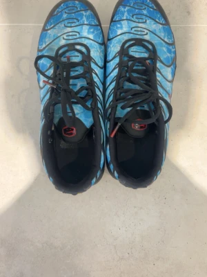 Nike Air Max Plus blå/svart sneakers - Nike tns  sneakers i blått med svarta vågmönster och röda detaljer. Skorna har en synlig Air-sula, svarta skosnören och en cool vatteninspirerad design på ovandelen. Perfekta för dig som vill sticka ut med din sneakerstil. Jag kan gå ner i priset 