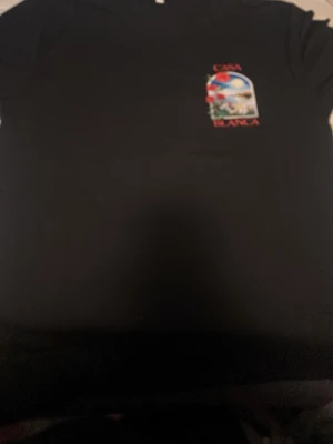 Svart Casa Blanca - Snygg svart t-shirt från Casa Blanca med stort färgglatt tryck framtill. Motivet visar ett fönster med röda rosor, blommor och en strand med solnedgång. Helt ny 