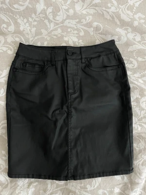 Svart kortkjol i skinn material  - Snygg svart kortkjol i skinnimitation med klassisk femficksdesign och bälteshällor. Kjolen har knapp och dragkedja framtill.