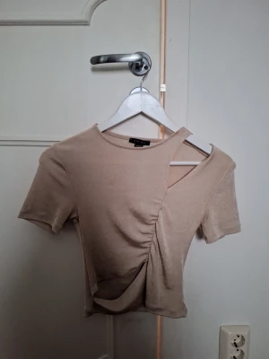 Beige topp  - En söt beige topp med ett passande cut out vid axeln. Kortärmad i mjukt material som sitter snyggt på kroppen. Perfekt för dig som vill sticka ut lite extra på fester. 