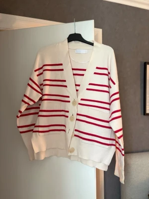 Vit kofta med röda ränder - Kofta i mjukt material med breda röda ränder på vit botten. Modellen har v-ringning, stora beige knappar framtill och ribbade muddar. Långärmad och avslappnad passform, perfekt för lager på lager och en chill look.