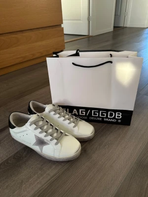 Golden Goose vita sneakers med stjärna - Snygga vita sneakers från Golden Goose med grå stjärna på sidan och svarta detaljer vid hälen. Skorna har grå skosnören och en klassisk låg siluett. Tillverkade i läder med slitna detaljer för en cool och avslappnad look.