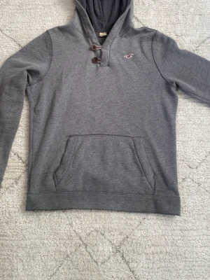 Grå Hollister hoodie med knappar - Snygg grå hoodie från Hollister med två knappar. Topp skick i storlek S. Hör av er vid minsta fundering!😀