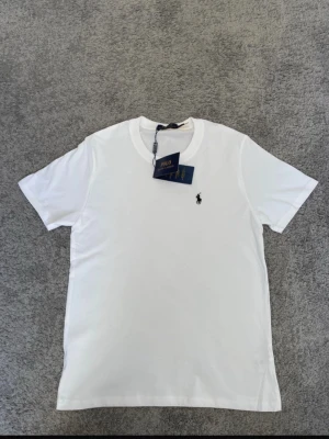 Vit t-shirt från Polo Ralph Lauren - Klassisk vit t-shirt från Polo Ralph Lauren med rund hals och korta ärmar. T-shirten har den ikoniska svarta broderade loggan på bröstet och är tillverkad i mjuk bomull för en skön känsla. Perfekt basplagg till garderoben.