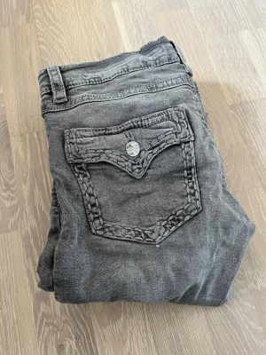 Grå jeans med flätad bakficka - Säljer ett par grå jeans från gina young med unik flätad detalj på bakfickan och knappstängning. Jeansen har en tvättad look och slitning vid benslutet för en cool, avslappnad stil. Perfekta för dig som gillar streetwear och vill sticka ut lite extra.