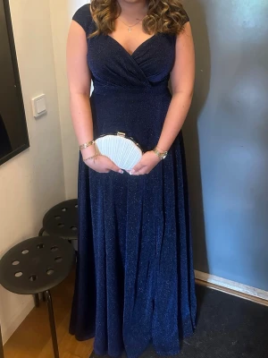 Marinblå glittrig ballklänning - Elegant marinblå långklänning med glittrigt tyg och v-ringad omlott överdel. Klänningen har breda axelband och en slits framtill som ger en snygg detalj. Perfekt för bal eller festliga tillfällen. Använd 2 gånger💙 orginal pris 1100kr
