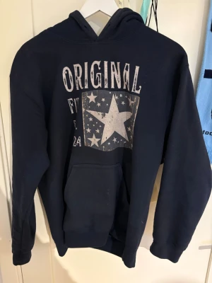 Mörkblå hoodie (original)  - Mörkblå hoodie med stor ficka framtill och huva. Tryck på bröstet med texten 'ORIGINAL' och en stor stjärna omgiven av mindre stjärnor. Skön och avslappnad passform, perfekt för chill dagar. Materialet känns mjukt och bekvämt.