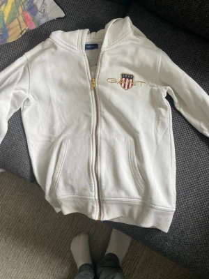 Vit hoodie med dragkedja från Gant - Säljer en vit hoodie från Gant med dragkedja framtill och fickor. Tröjan har huva och ett broderat Gant-emblem med sköld och texten 1949 på bröstet. Tillverkad i mjukt bomullsmaterial och har ribbade muddar vid ärmslut och nederkant.