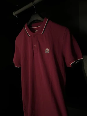 Lila pikétröja från Moncler - Snygg Lila pikétröja från Moncler med klassisk krage och knappar framtill. Tröjan har vita och marinblå detaljer på krage och ärmslut samt Moncler-logga på bröstet. Tillverkad i mjuk bomull och har korta ärmar.