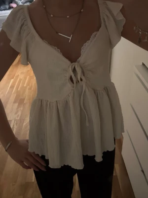 Vit topp med volang från 157 - Supersöt vit topp från 157 med volangdetaljer och knyt framtill. Toppen har korta ärmar och v-ringning. Perfekt till jeans eller kjol för en fräsch look i sommar.