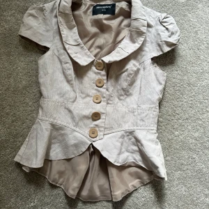 Ljusrosa blus med volang och rosett - Supersöt beige blus från Atmosphere med stora knappar framtill, volang nertill och en fin rosett i ryggen. Blusen har korta puffärmar, markerad midja och bred krage. 