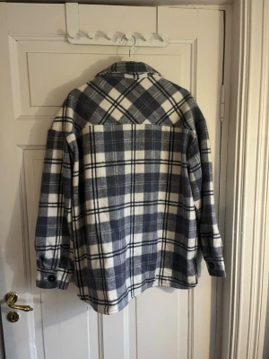 Rutig overshirt i blått och vitt - Overshirt i mjukt,  rutigt mönster i blått och vitt. Två bröstfickor med knappar, sidfickor och klassisk krage. Stängs med knappar framtill och har knappdetaljer vid ärmsluten. Perfekt lager på lager-plagg för kyligare dagar.