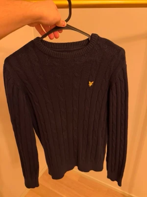 Mörkblå kabelstickad tröja Lyle & Scott - Mörkblå kabelstickad tröja från Lyle & Scott med rund halsringning och gul broderad logotyp på bröstet. Tröjan har långa ärmar och ribbade muddar vid ärmslut och nederkant. Perfekt för dig som gillar klassisk och stilren look.
