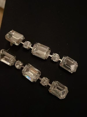 Glittrande örhängen med stora kristaller - Säljer ett par eleganta örhängen med stora, rektangulära och runda klara kristaller. Örhängena har en silverfärgad infattning och ger ett riktigt lyxigt intryck. Perfekt för dig som gillar att sticka ut och vill ha något extra till din look.