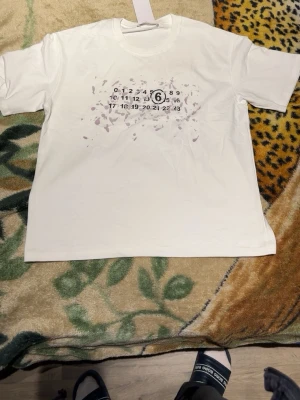Vit Maison Margiela t-shirt - Säljer en vit t-shirt från Maison Margiela med det ikoniska siffertrycket och diskreta färgstänk på bröstet. T-shirten har rund hals och korta ärmar. Perfekt för dig som gillar minimalistisk design med en twist.